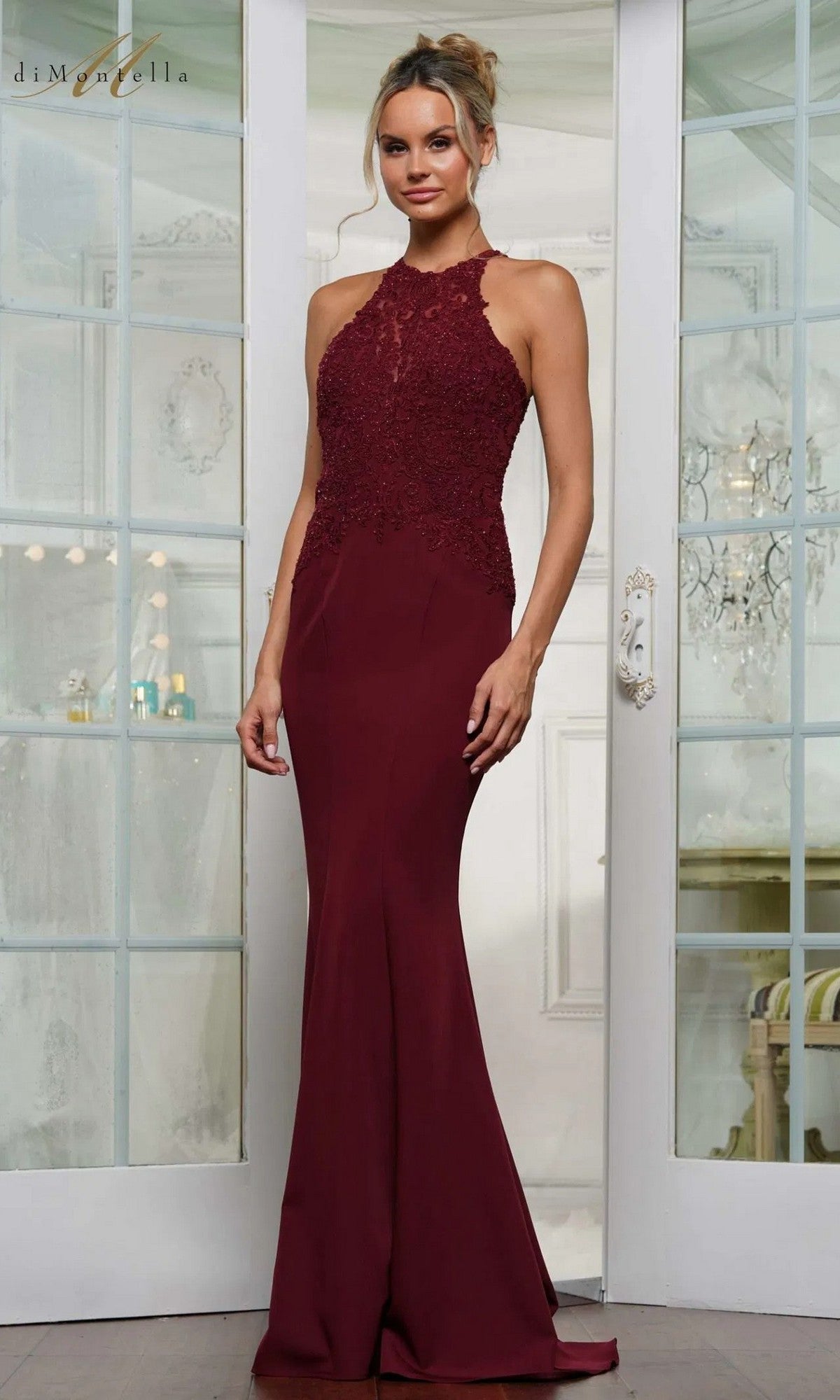 A woman stands indoors wearing the Rina Di Montella RD3243 long formal dress in wine, featuring a sleeveless halter neck, floor-length crepe design, and a lace-embroidered bodice.